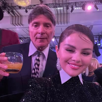 Two people posing for a photo at an event; one holding a drink, both dressed in formal attire with a blurred background.