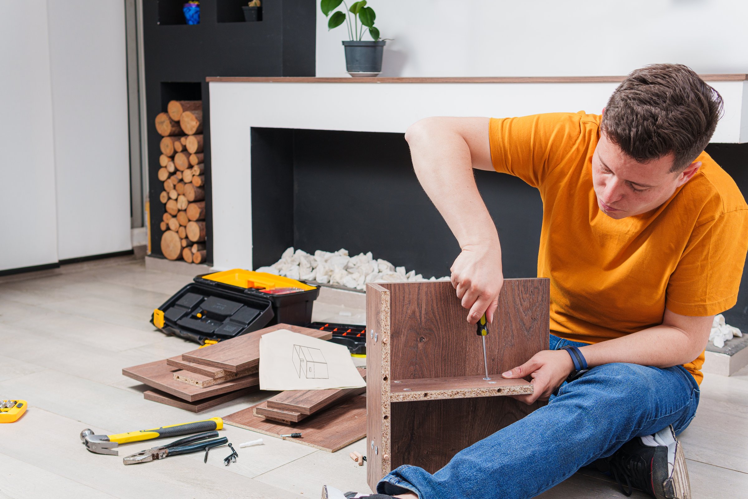 A young hardworking man is assembling a piece of furniture in the living room of an apartment. He is screwing a piece of wood and there are several tools on the floor like a hammer and pliers