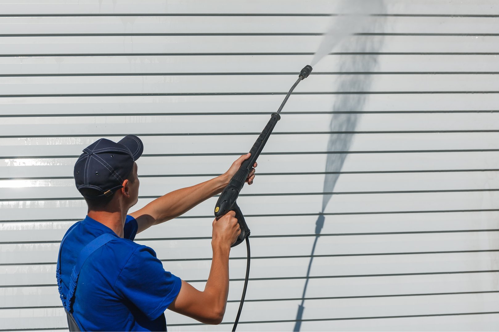 Worker in overalls washes a white wall from a siding with a water gun. Cleaning service for washing buildings and facades