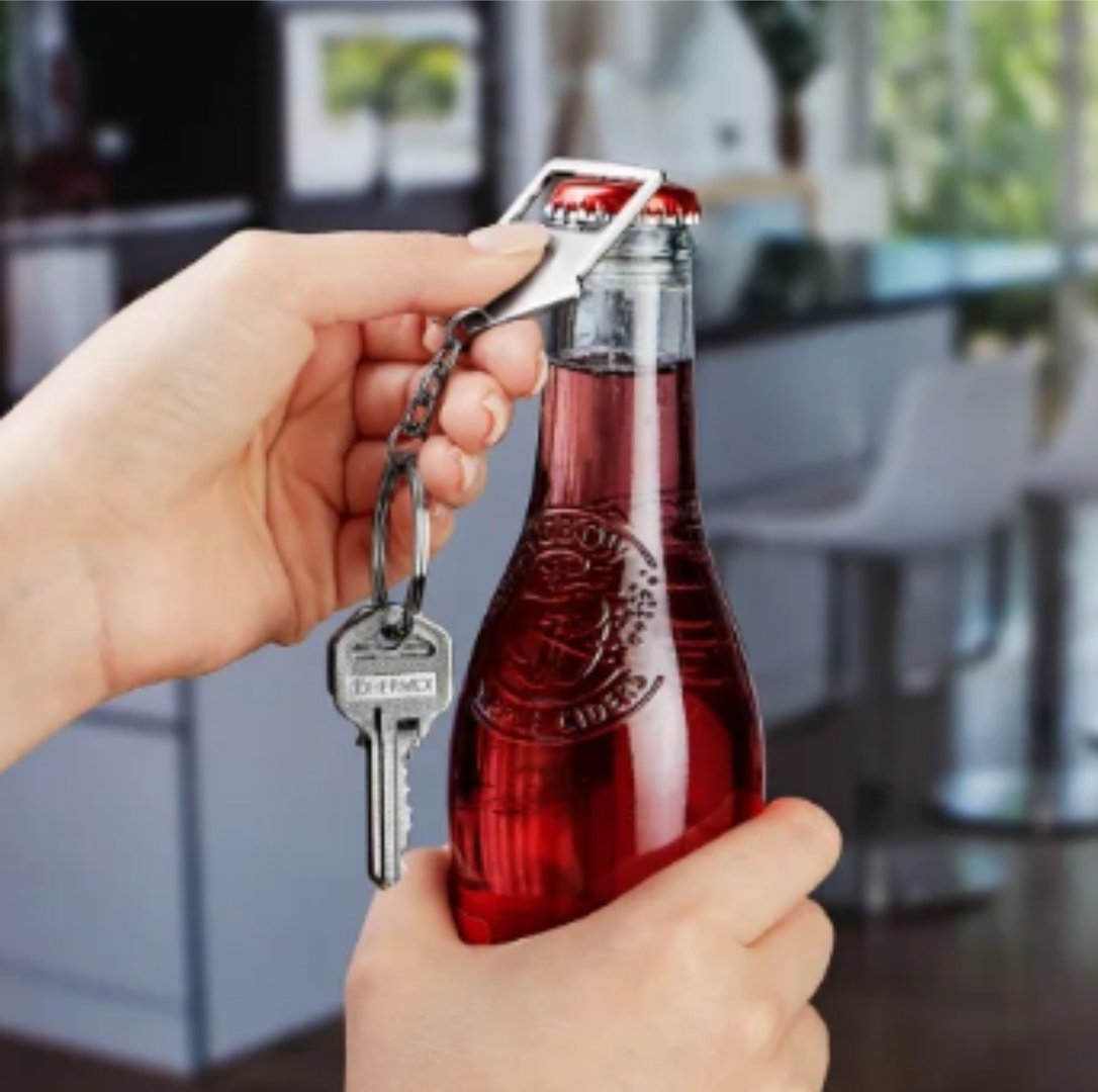 Hand using a keychain bottle opener to open a glass bottle of red liquid in a modern kitchen setting.