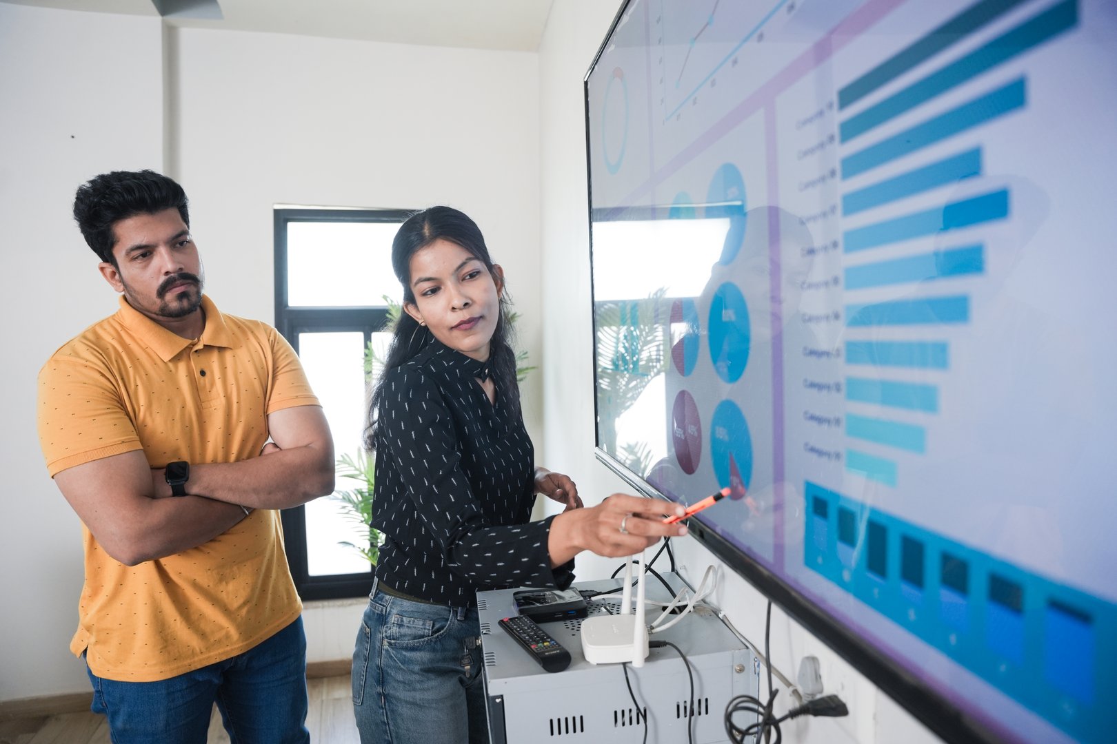 Young indian woman explaining company growth strategy through presentation to colleague on wall tv in office. analysis, statistics charts and graph concept.