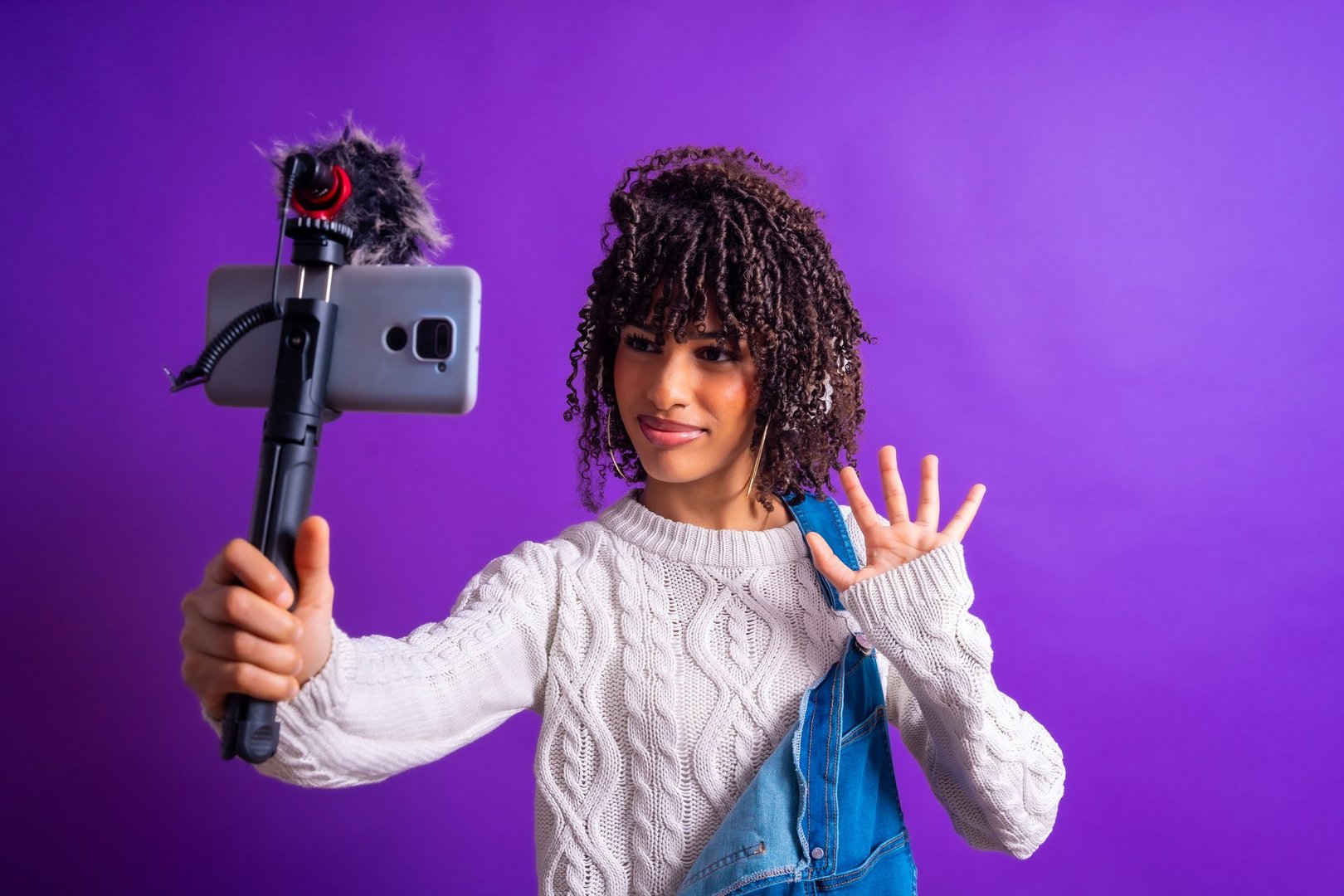 Young female vlogger waving and smiling while recording a video blog using her smartphone on a tripod with external microphone