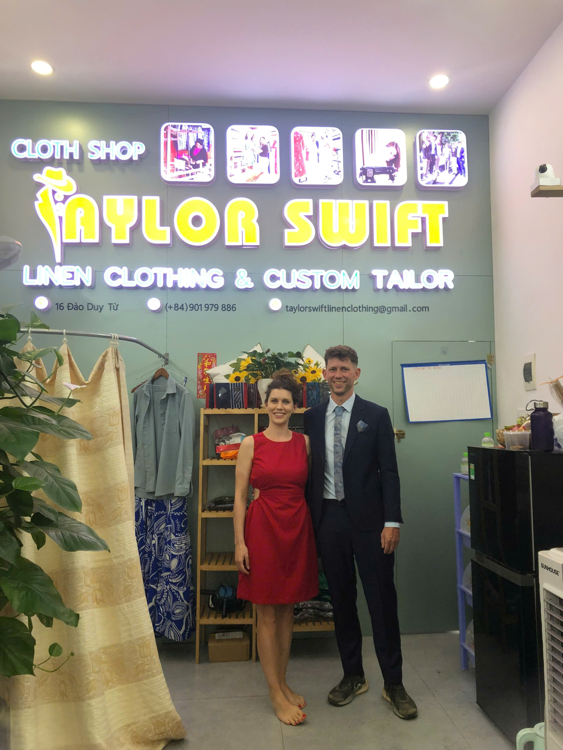 Couple posing inside a tailor shop with a "Taylor Swift" sign, offering linen clothing and custom tailoring services.