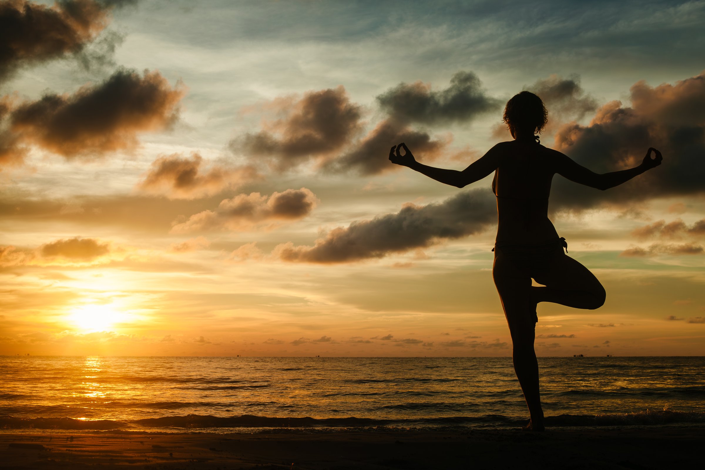 A woman practicing yoga while standing on a tropical beach during a stunning sunset, surrounded by serene beauty and warm light.