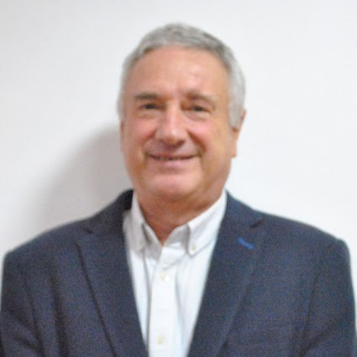 A smiling man with gray hair wearing a dark suit jacket and white collared shirt, standing against a plain white background.