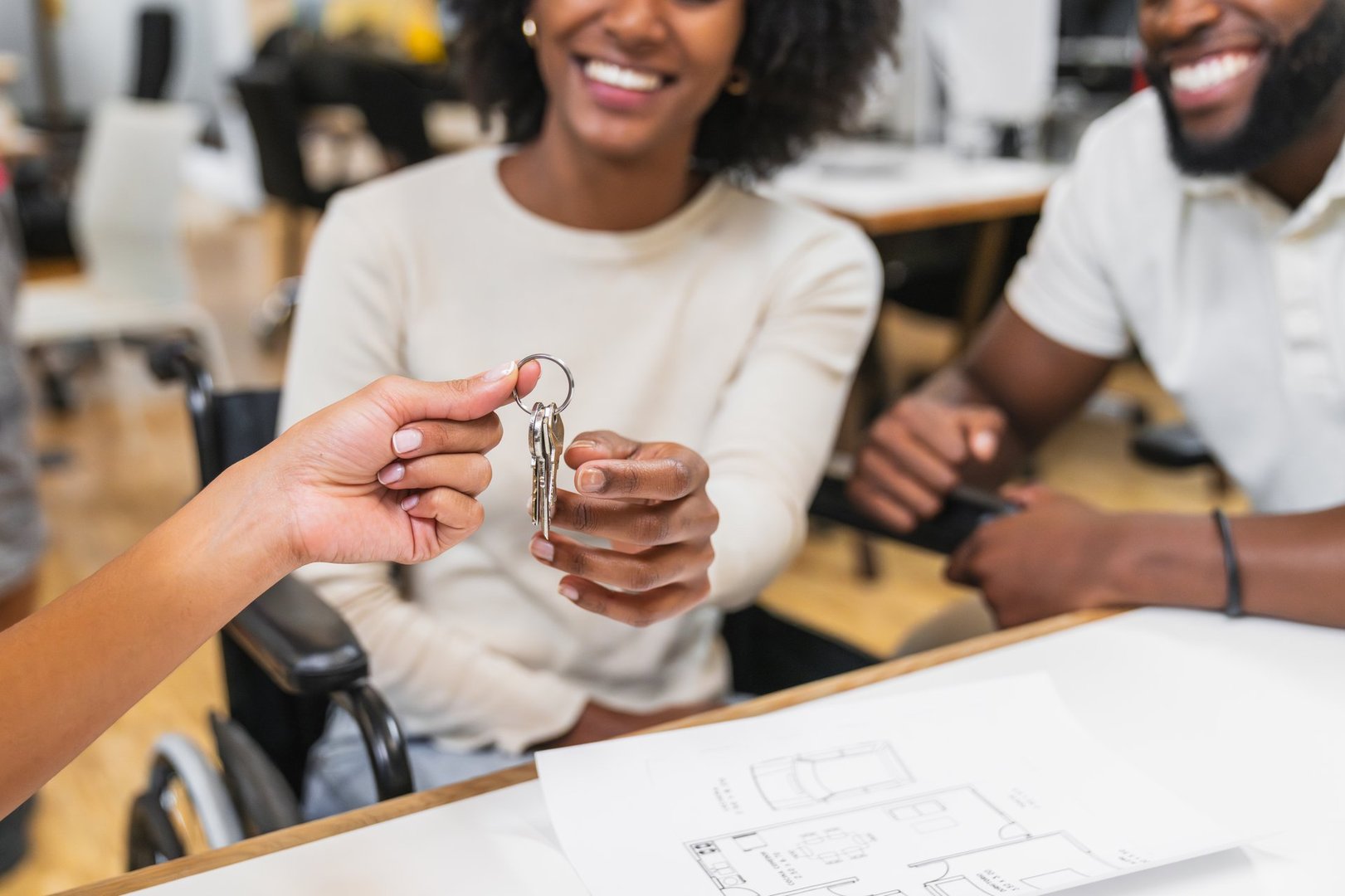 Real estate agent handing over new house keys to a happy young couple in a coworking, blueprints are visible on the table