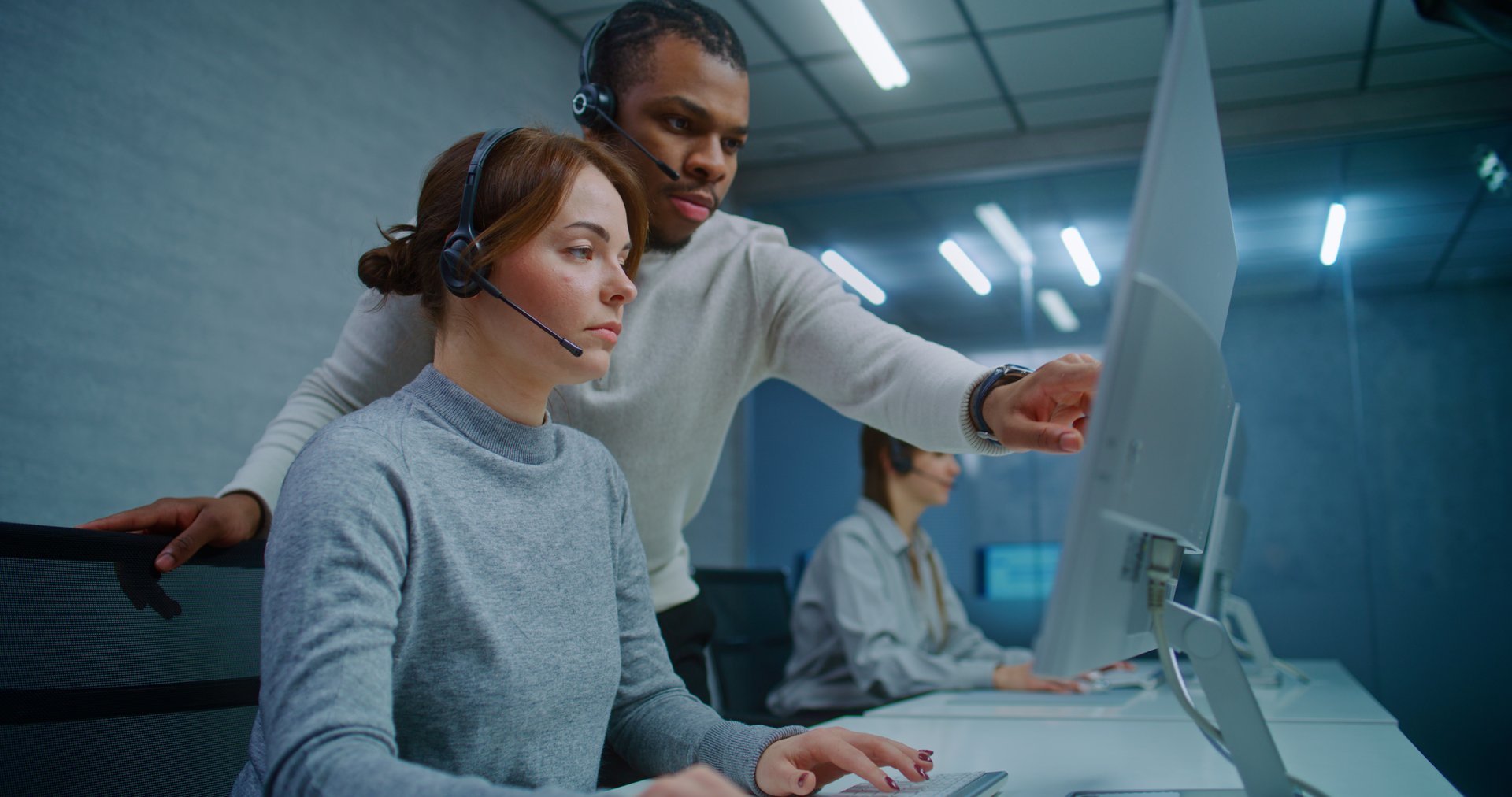 Call center office: Female technical support specialist in headset monitors data on computer with African American colleague. Diverse team of hotline operators work in online customer support service.