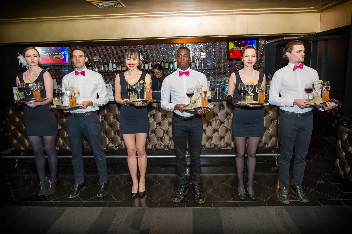 Group of waitstaff in uniform holding trays with drinks, standing in a line in a restaurant or bar setting.