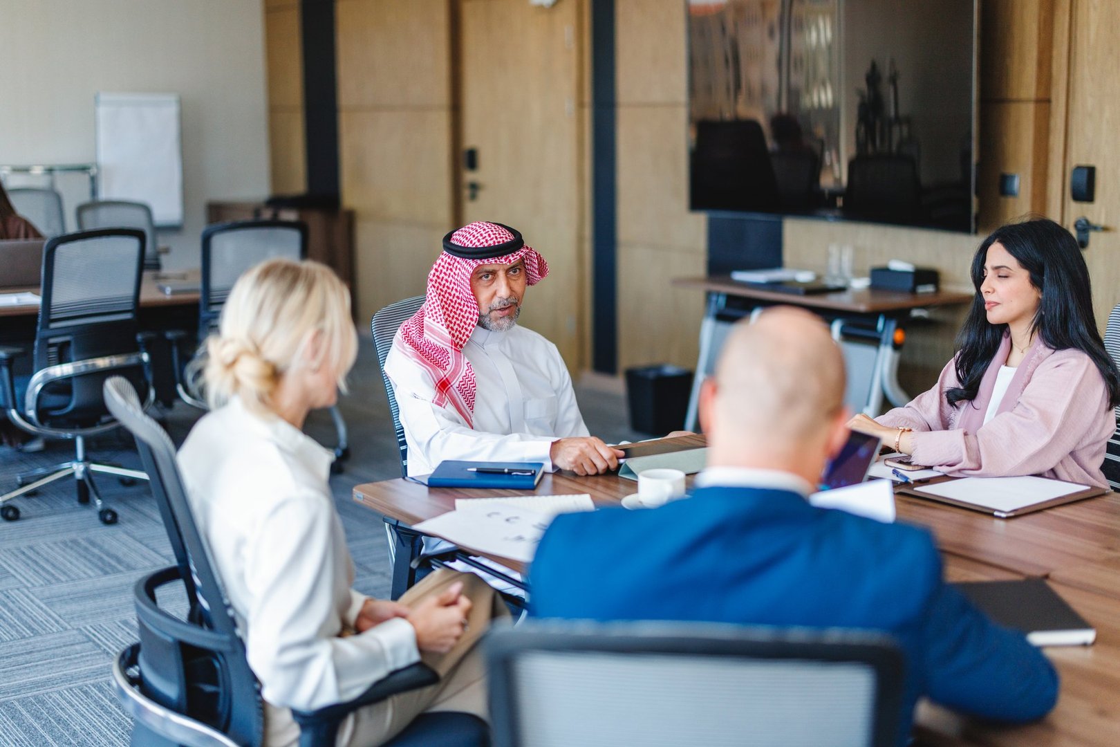 Middle Eastern business executives engaged in a focused discussion in a modern office. The group appears involved and attentive, sitting around a table with documents and notepads, featuring a corporate environment with office chairs and a large screen.