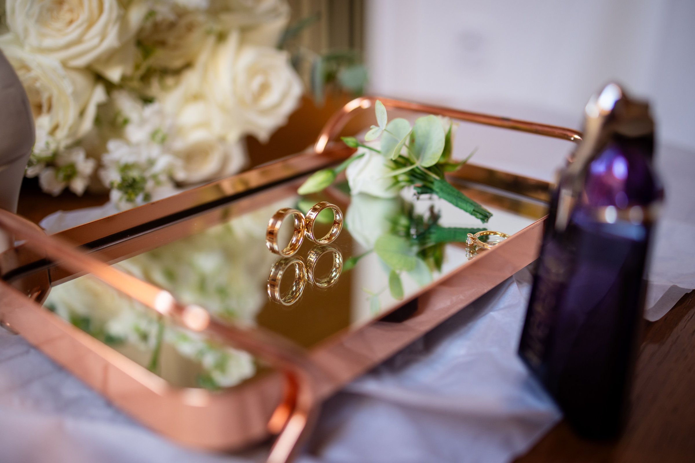 Elegant gold wedding rings on a mirrored tray with white roses and a perfume bottle, symbolizing romance and celebration.