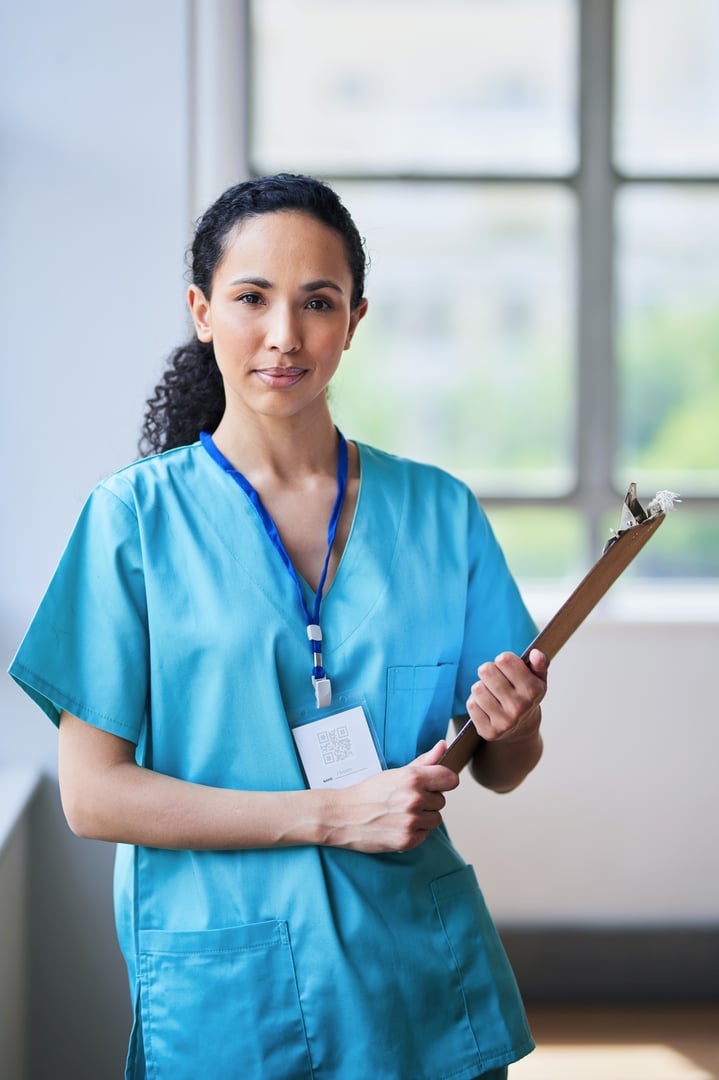 A confident female nurse, dressed in blue scrubs, stands in a hospital corridor holding a clipboard. She is prepared and ready for her shift, symbolizing professionalism and dedication in healthcare.