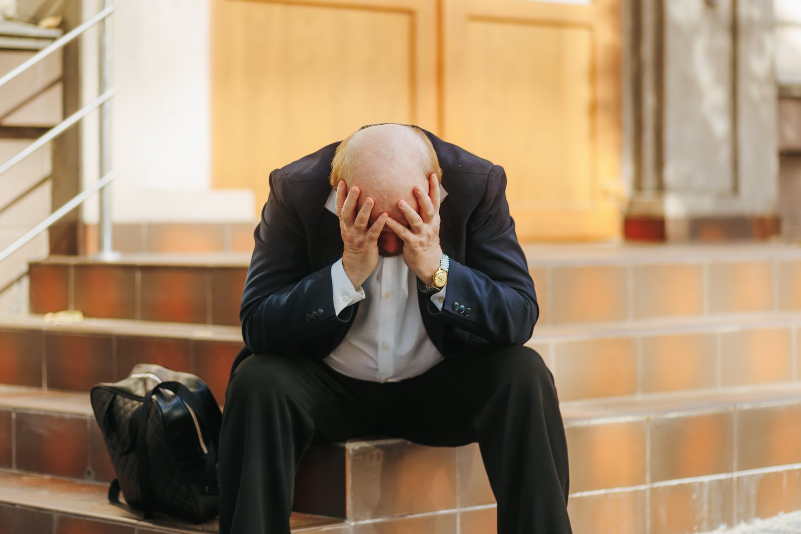 A senior bald office worker sits outdoors on stair steps, face in hands, feeling exhausted and emotionally drained, symbolizing burnout, stress, and despair.