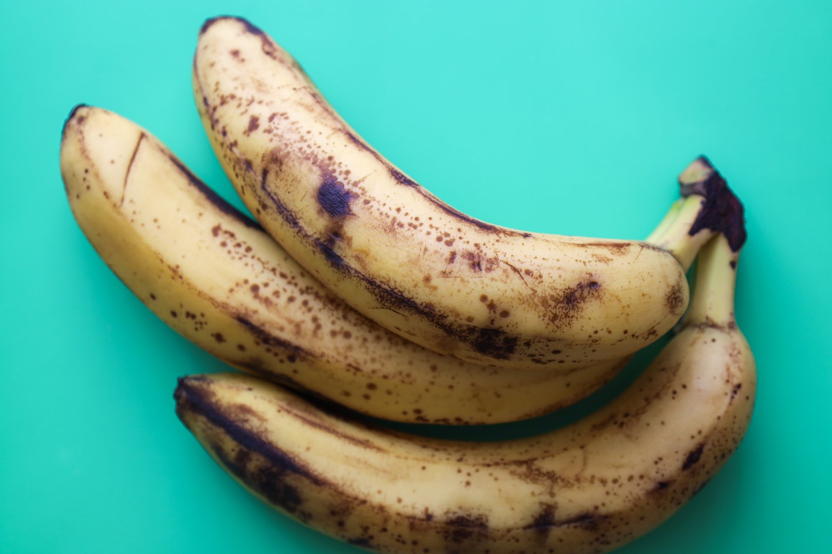 Three ripe bananas with brown spots rest on a vibrant turquoise background, creating a contrast of colors and textures. Sweet tropical fruits, ready for eating or baking.