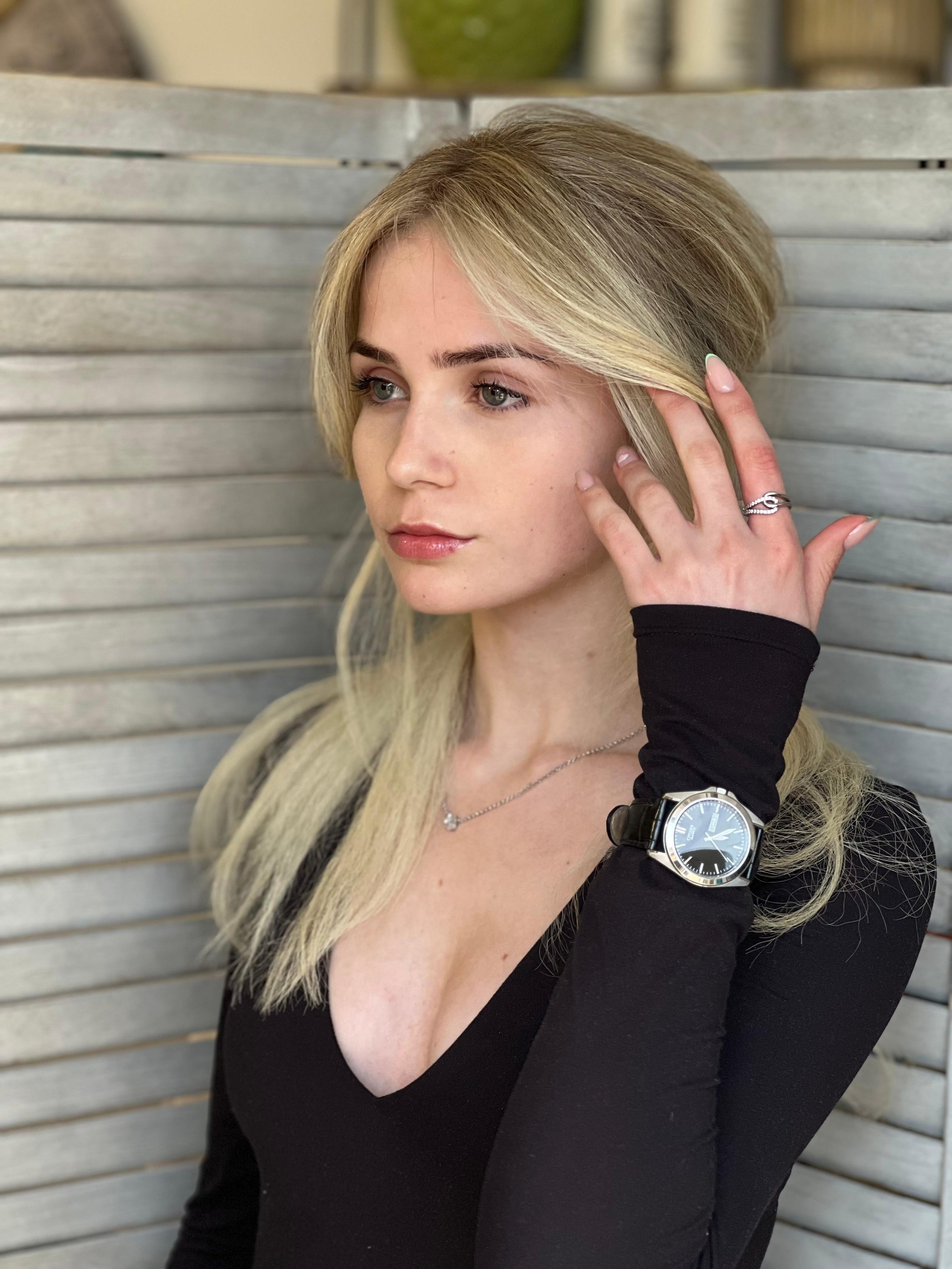 Woman with long blonde hair wearing a black top and watch, standing in front of a wooden panel, touching her hair.