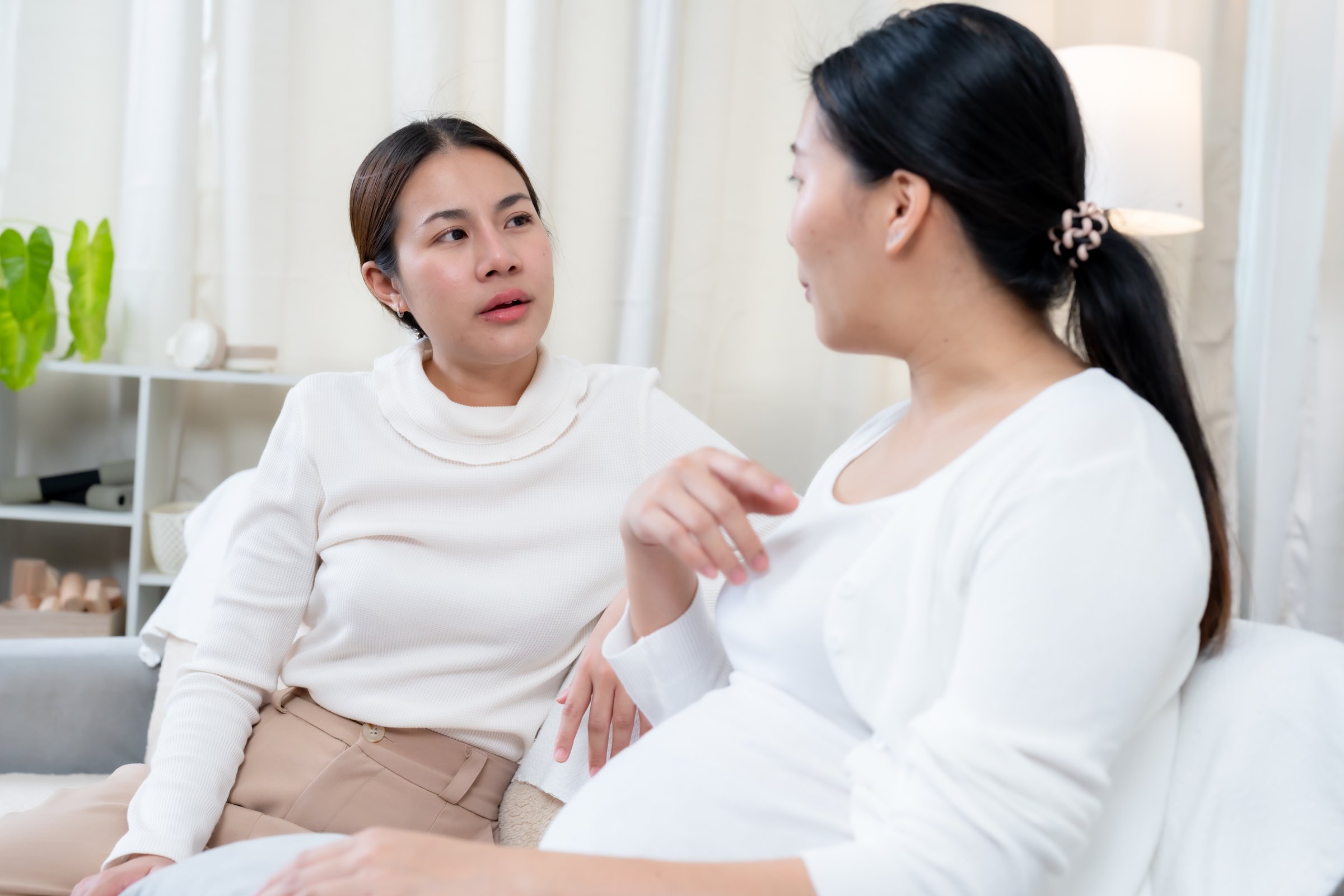 Beautiful pregnant Asian woman nearing birth. Engaged in deep conversation with a close friend, sharing concerns and emotions. Warm connection, care, and support in a cozy home setting.