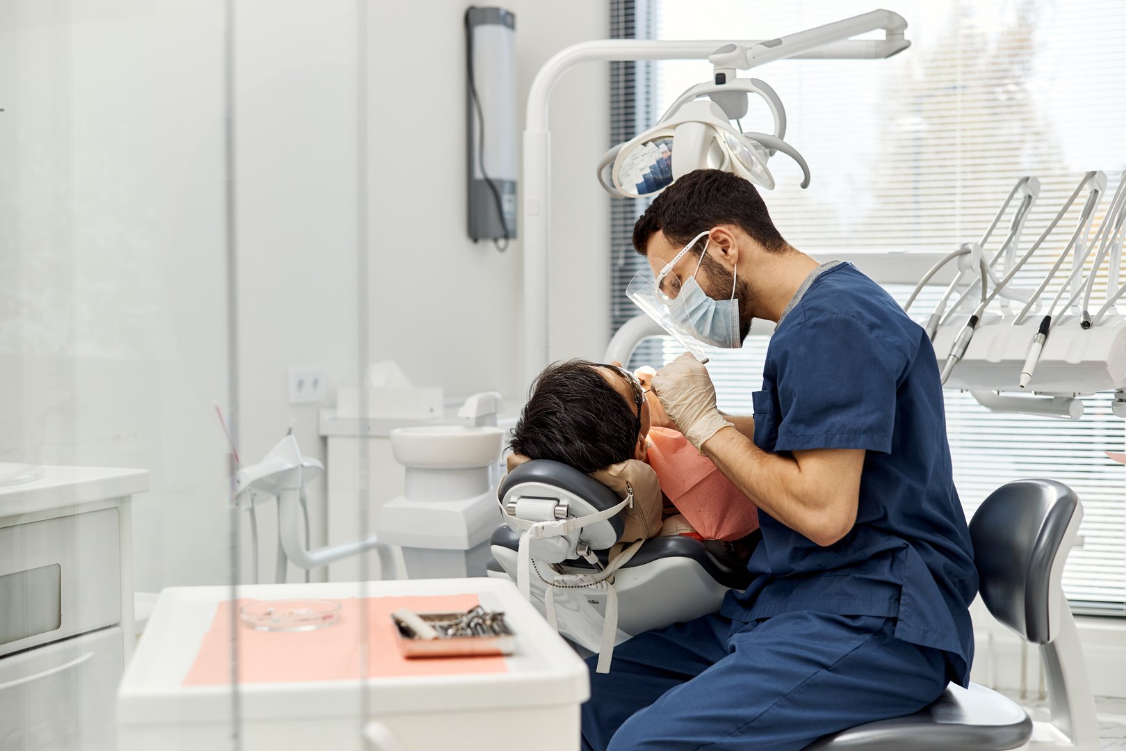 Middle Eastern male dentist examining patient in dental chair during routine checkup in modern dental office, both wearing protective masks, dental tools visible in foreground