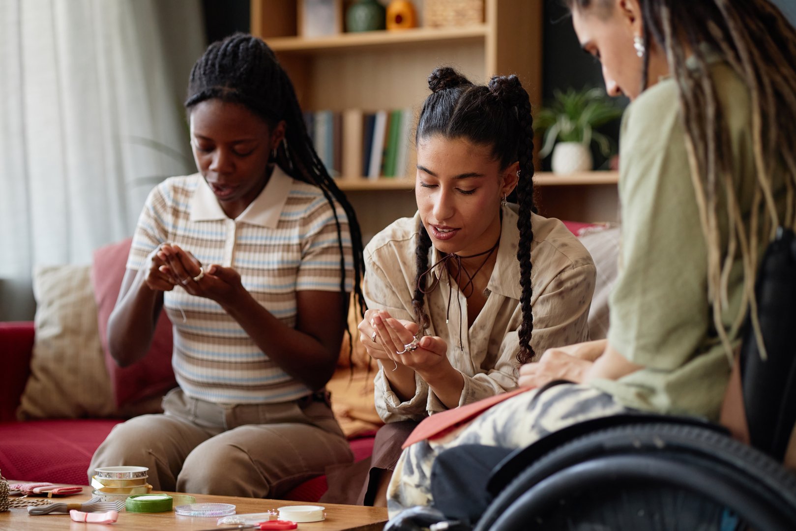 Three young diverse girlfriends including one with disability engaging in creative activity, making handmade jewelry at home