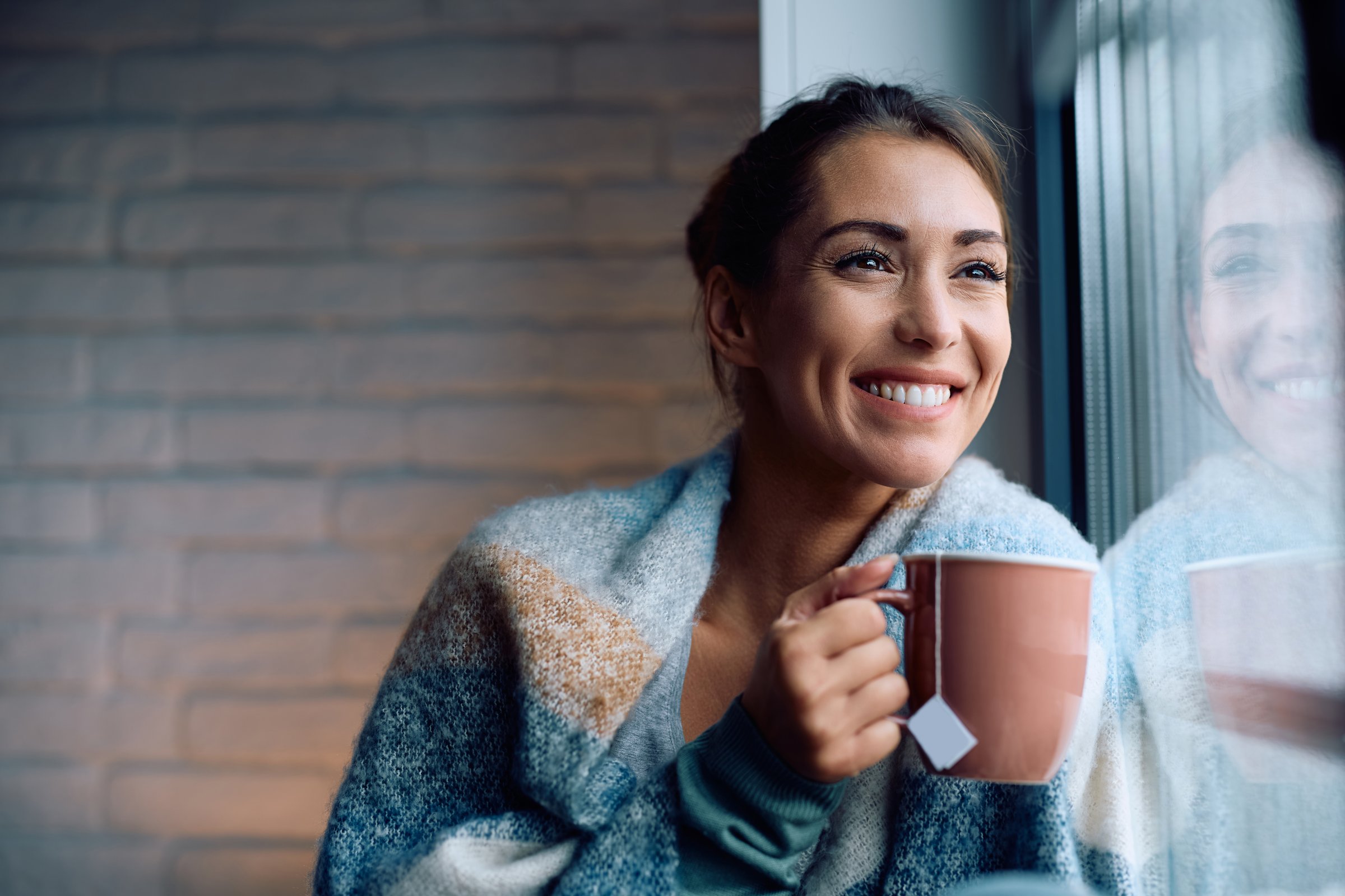 Smiling woman drinking tea while relaxing by the window and looking through it. Copy space.