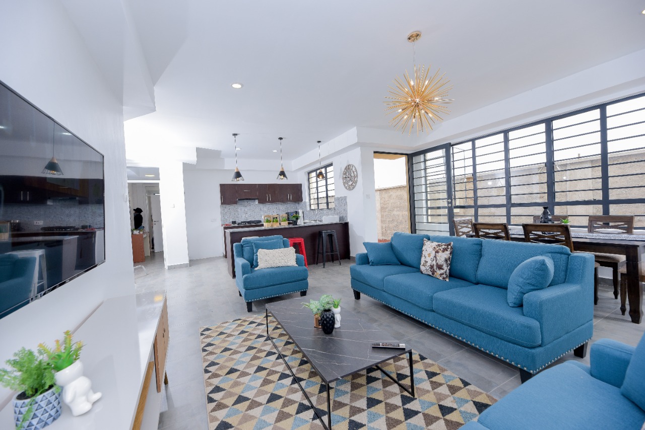 Modern living room with blue sofas, geometric rug, TV, and open-plan kitchen with pendant lights and dining area.