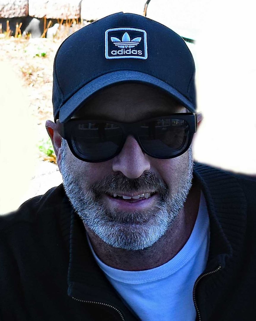 Man with a black Adidas cap and sunglasses smiling outdoors. He has a beard and is wearing a white shirt and dark jacket.