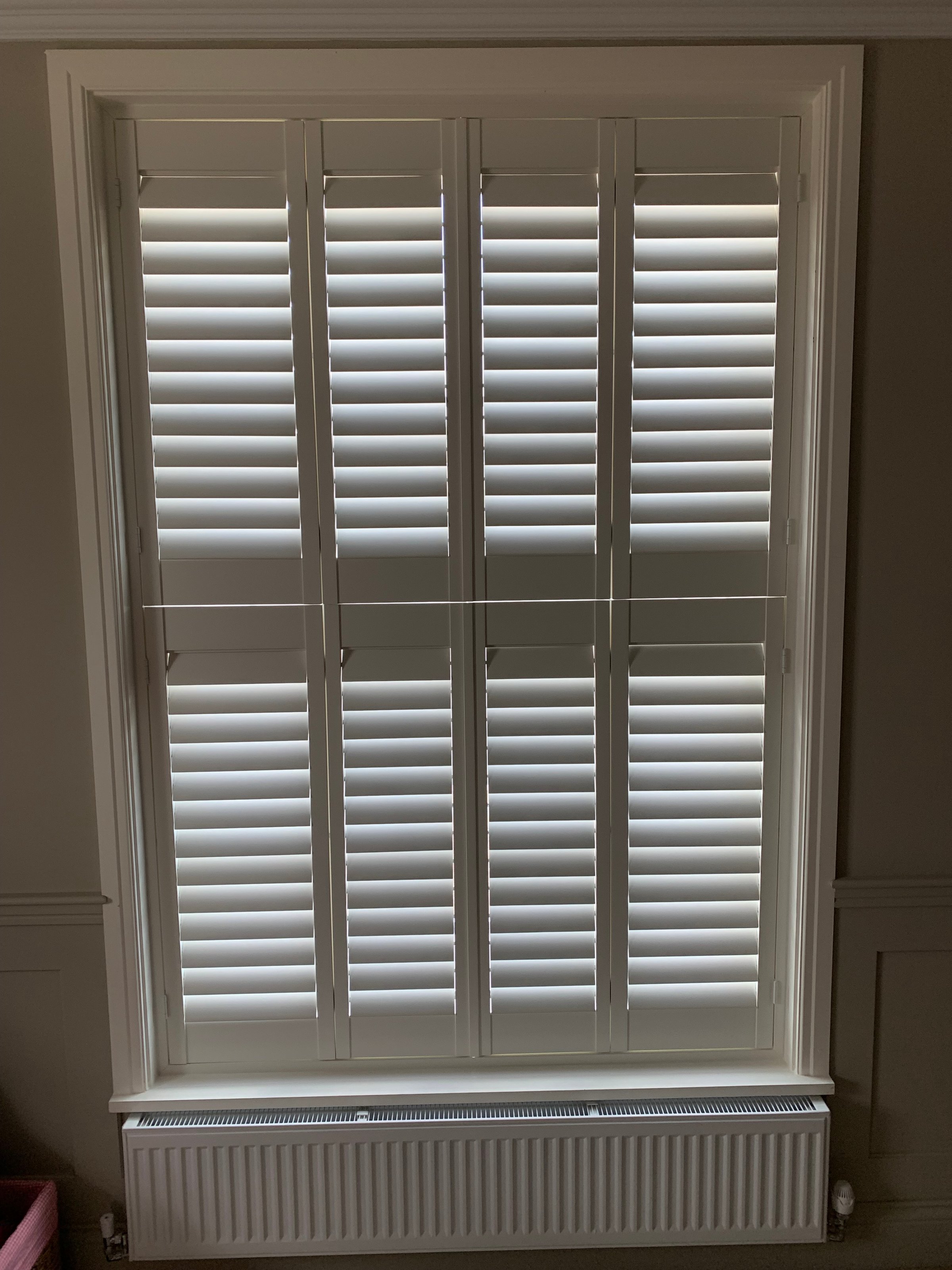 A window with closed white plantation shutters, diffusing light into a dimly lit room with a plain wall and a radiator below.