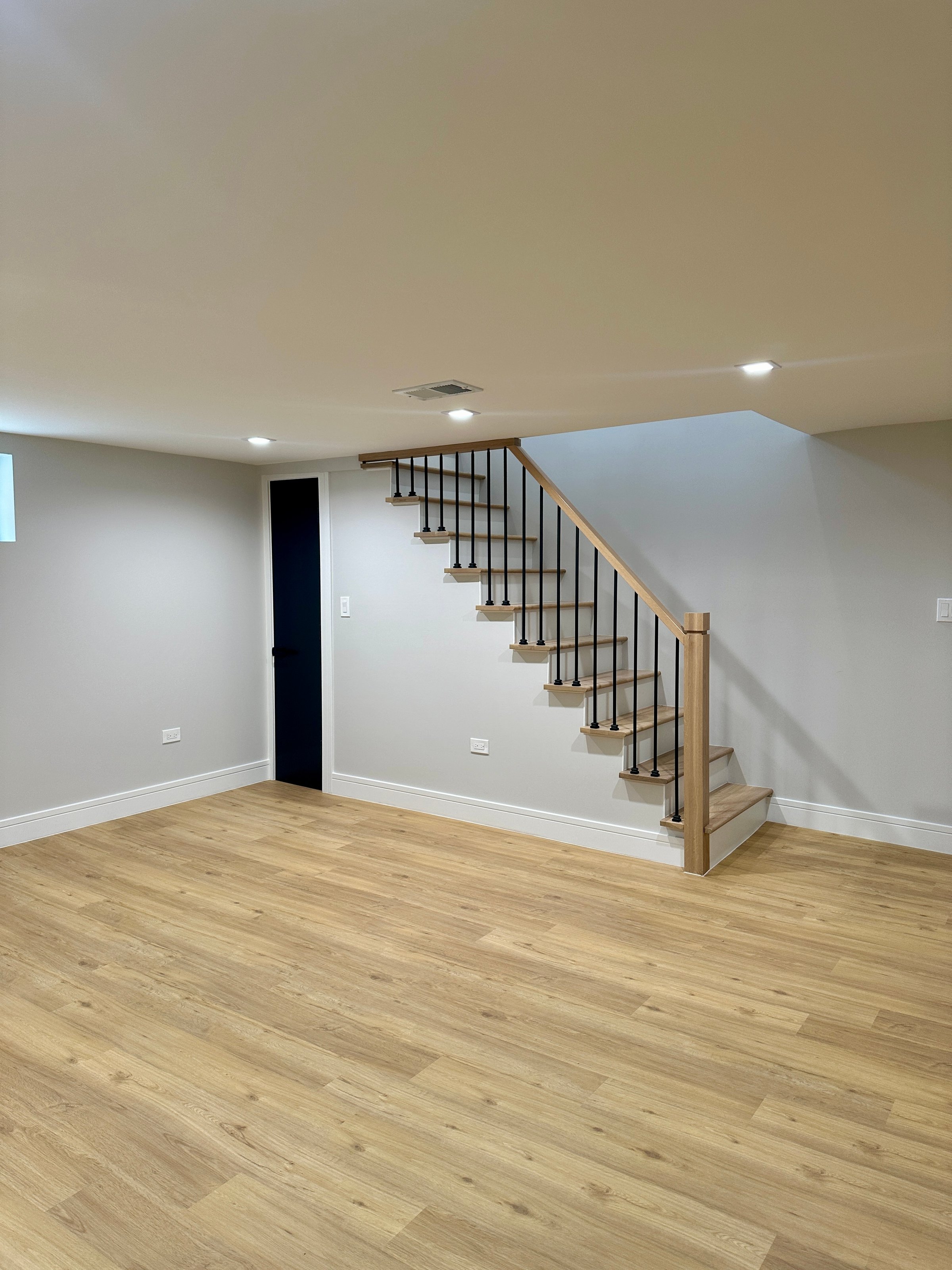 Empty basement room with light wood flooring, white walls, and a staircase with wooden banister and black balusters.