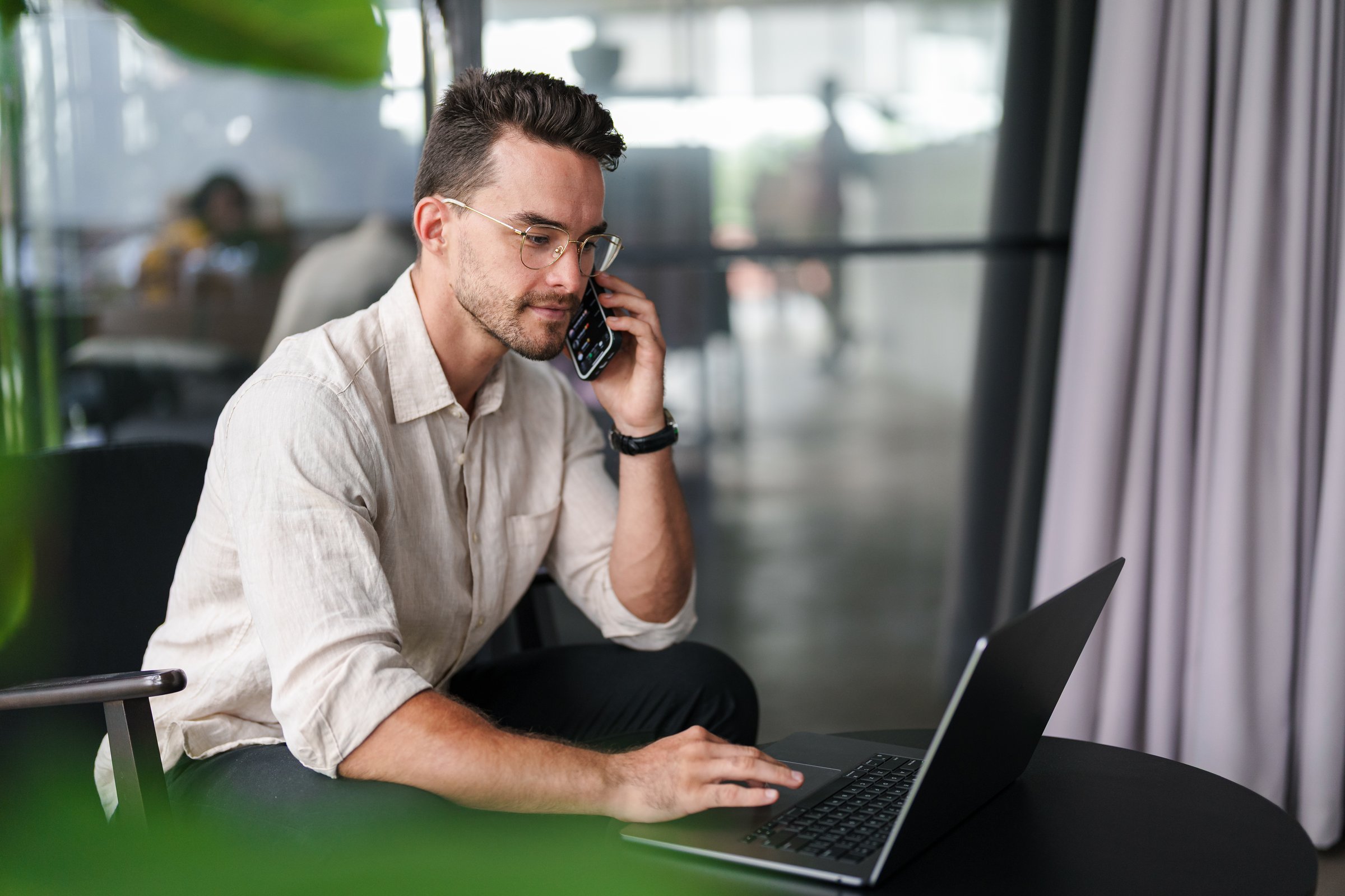 A young man with glasses, wearing a beige linen shirt, works on a laptop while speaking on the phone in a modern office setting.