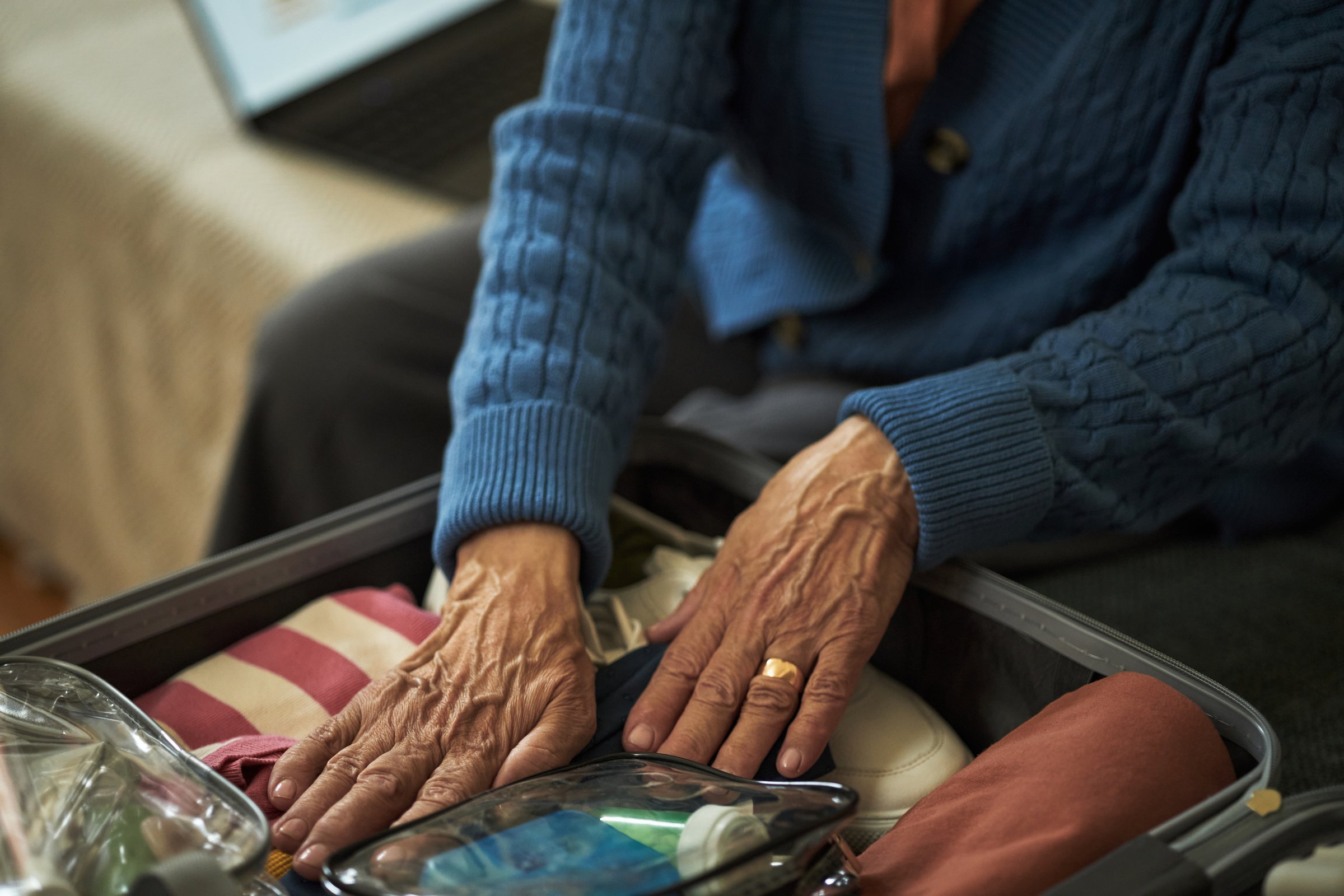 Elderly woman with a sweater packing suitcase, organizing belongings including clothes and toiletries. Hands prominently featured, demonstrating careful preparation