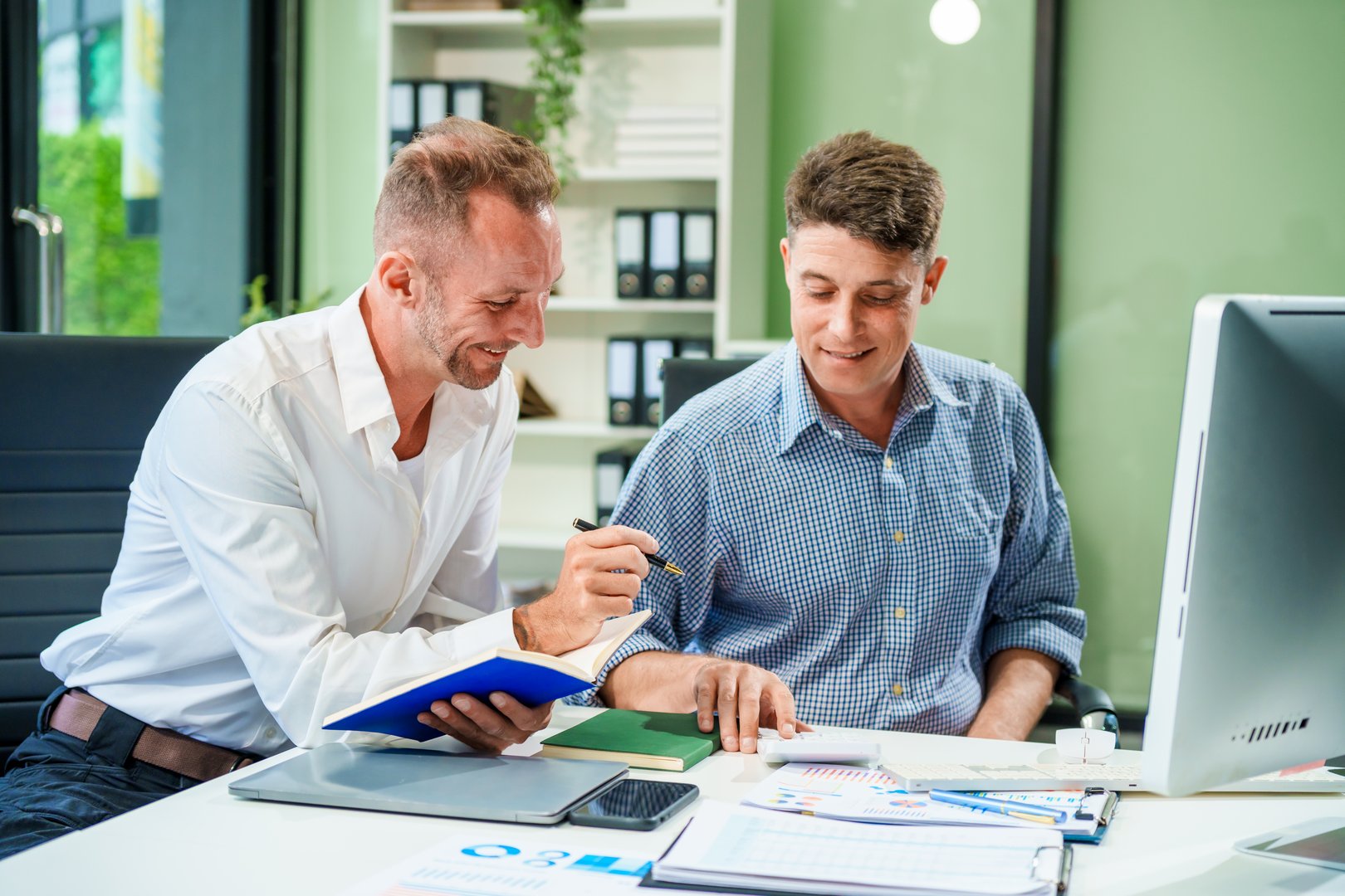 An Italian male and a Caucasian male sit at business desk in office meeting room, discussing financial terms, investment strategies, managing funds with computer monitor displaying relevant data.