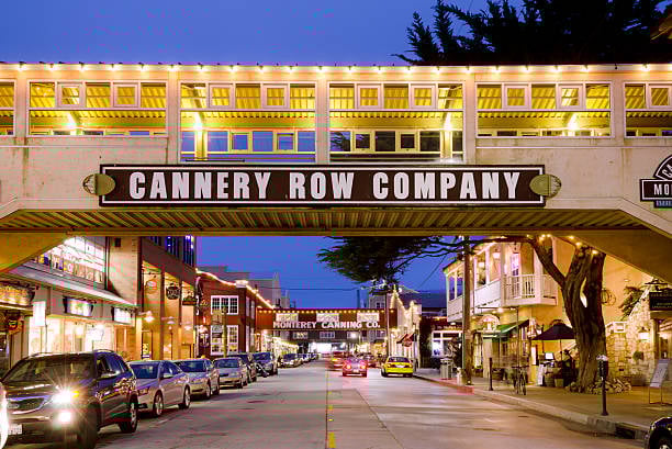 Evening view of Cannery Row, Monterey, featuring a lit pedestrian bridge and lined with shops and parked cars.