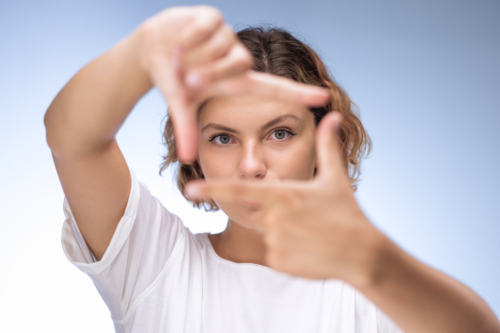 A determined young woman with short curly hair, using her hands to frame her face against a light background. Perfect for concepts of creativity, determination, and focus. High-quality stock photo ideal for various promotional and advertising materials.