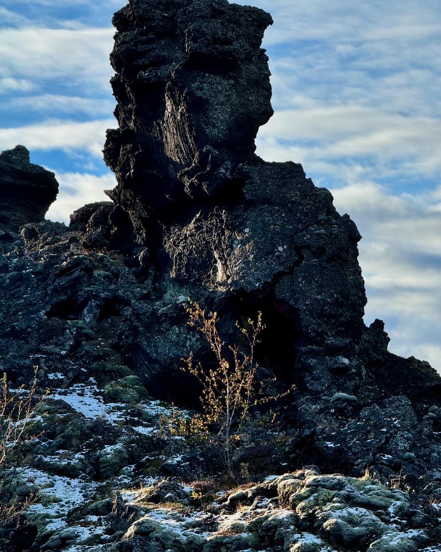 A rugged rock formation with a prominent, tall rock under a cloudy sky, surrounded by sparse vegetation and light snow patches.