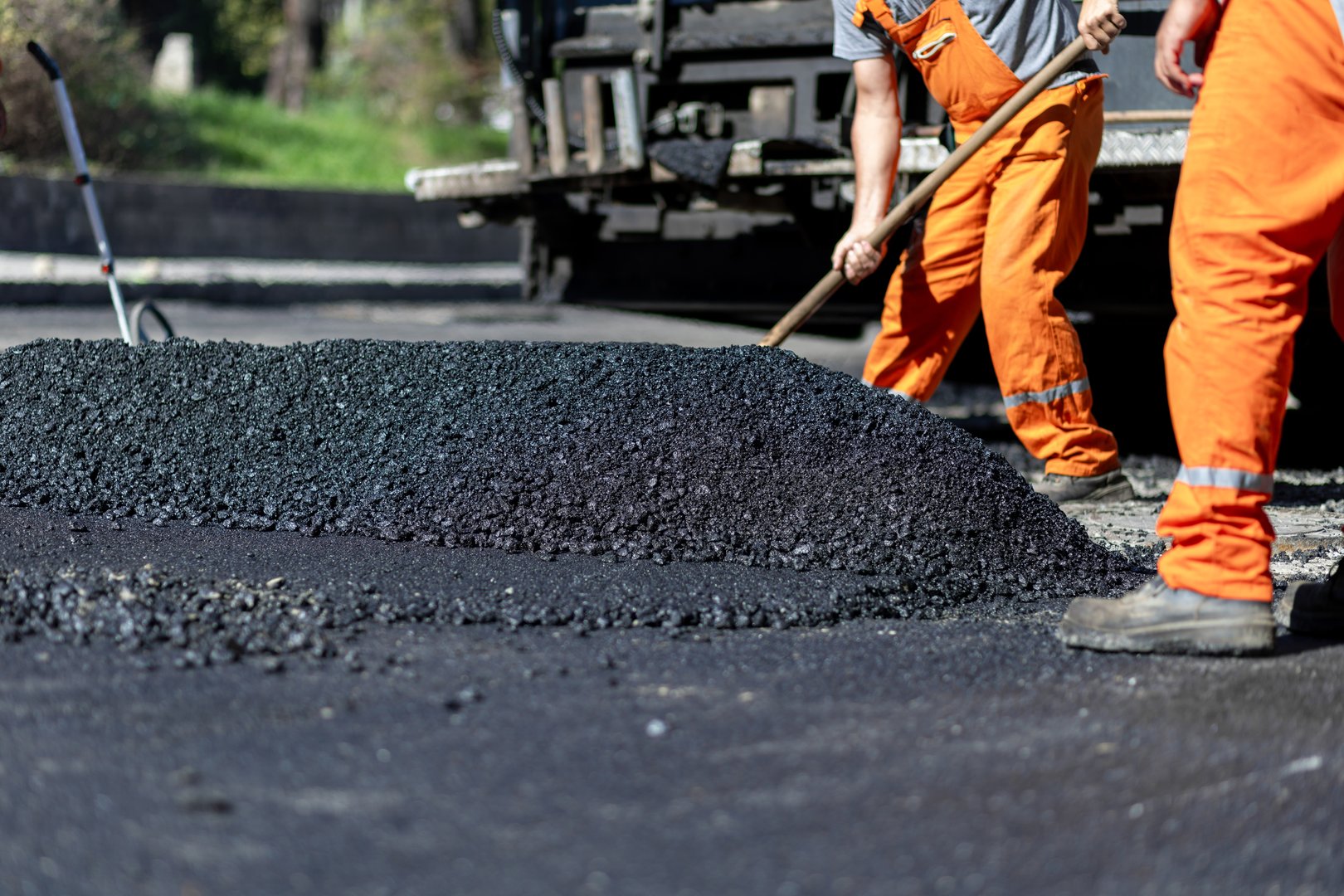 Construction workers in orange overalls are actively spreading hot asphalt on a road, ensuring a level finish for improved traffic flow and safety.