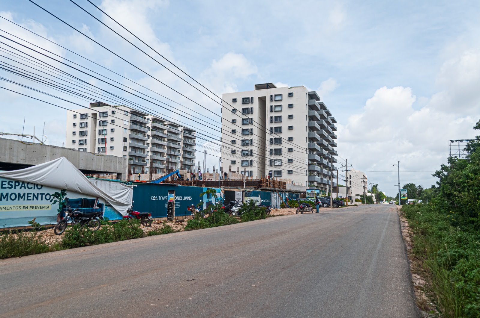 Construction site with cranes and unfinished buildings beside a road under a cloudy sky.