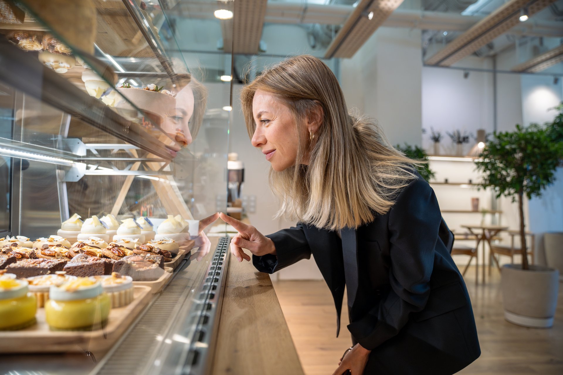 Happy woman looking at snacks for coffee, standing at showcase in cozy small cafe, going to buy purchase lunch, breakfast. Satisfied cheerful businesswoman at window display in coffeehouse, bakehouse.