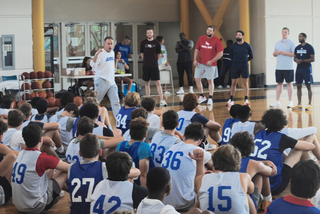Coach speaking to a group of young basketball players seated on a gym floor, with other staff standing in the background.