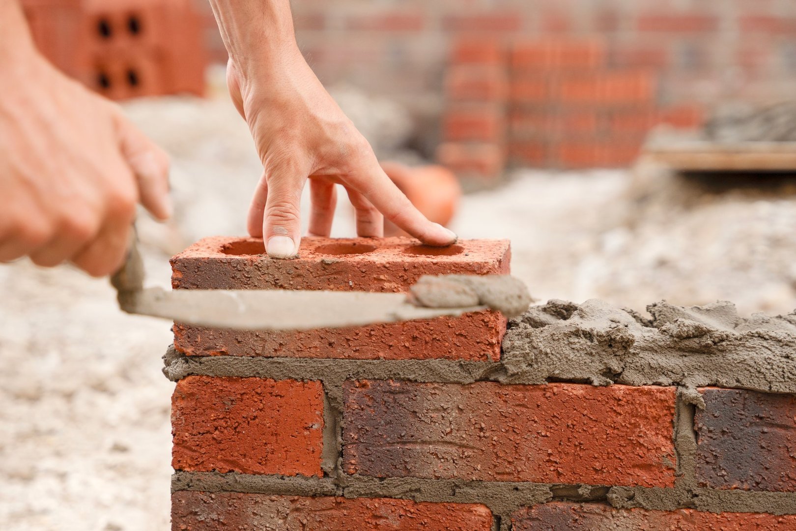 A worker is carefully placing bricks with precise movement on a construction site, showcasing skilled masonry work amid a developing structure.