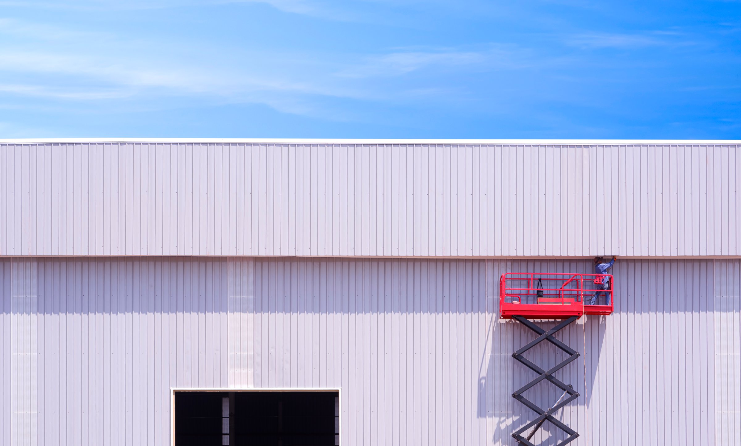 Construction worker on hydraulic scaffolding are installing corrugated steel wall on factory building structure in construction site against blue sky background