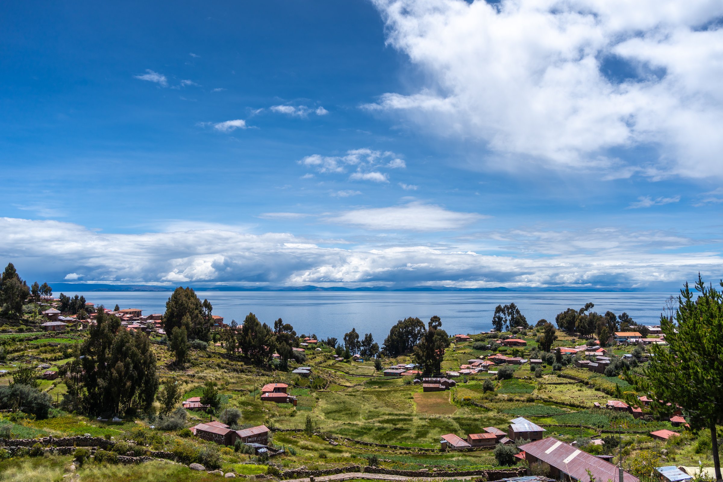 Traditional houses with red roofs and cultivated fields are spread across the green landscape of Taquile Island, overlooking the deep blue waters of Lake Titicaca in Peru. A peaceful rural setting