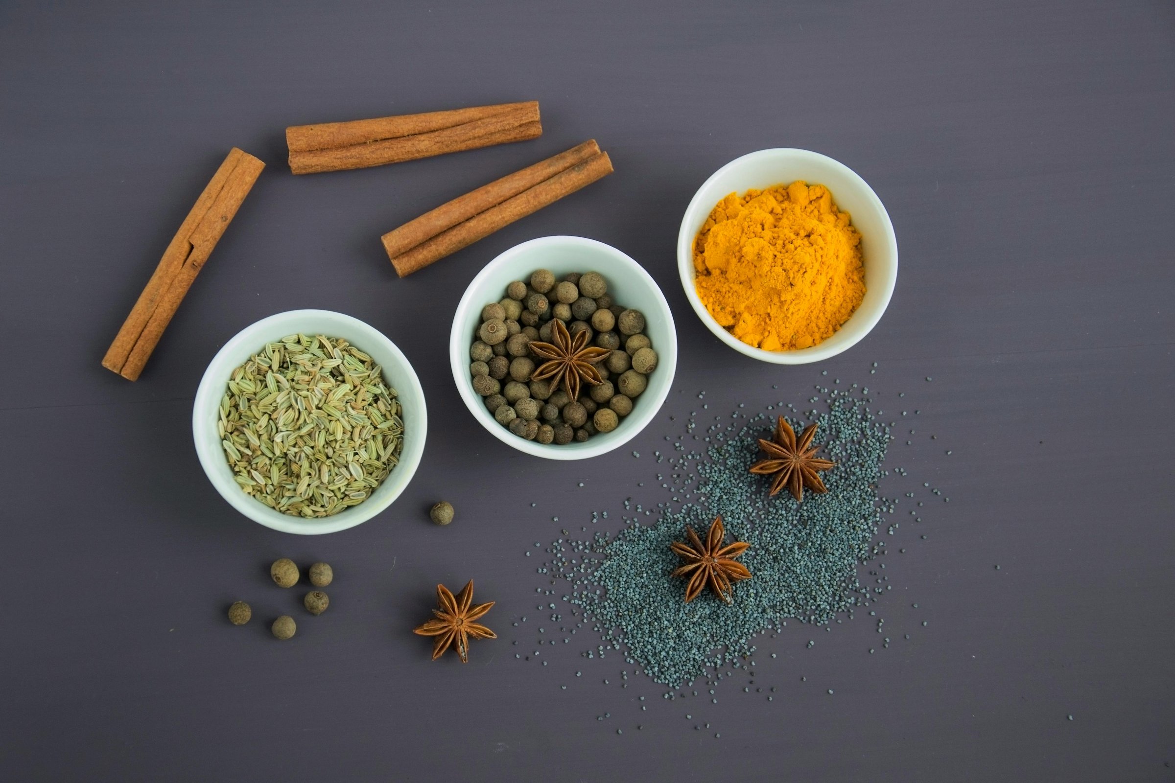 Spices in bowls: turmeric, allspice, fennel seeds, with star anise, cinnamon sticks, and poppy seeds on a gray background.