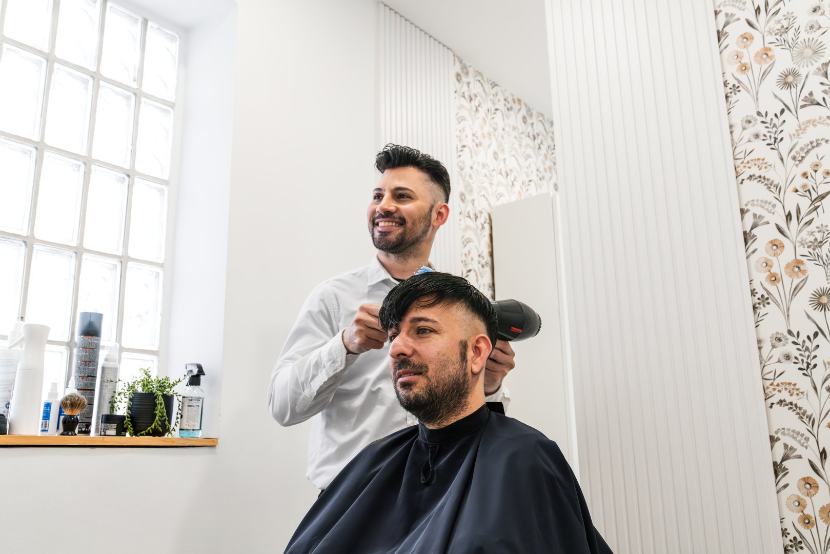 Hairdresser styling hair of a male customer using a hair dryer and comb in a contemporary hair salon, creating a fresh look
