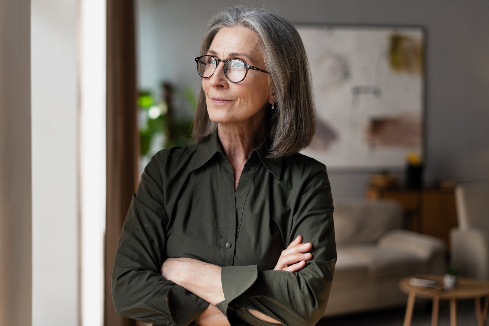 Mature businesswoman with glasses stands confidently with her arms crossed, gazing thoughtfully into the distance