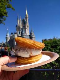 A hand holding a churro ice cream sandwich with a castle in the blurred background against a clear blue sky.