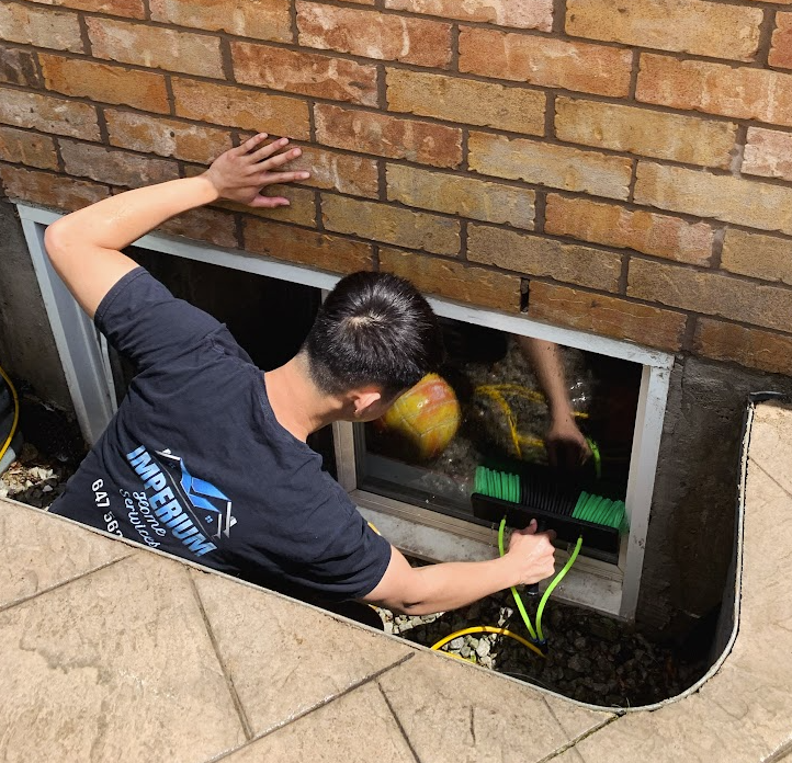 Person cleaning a basement window from outside, using a brush attached to a hose, surrounded by a brick wall and patio.
