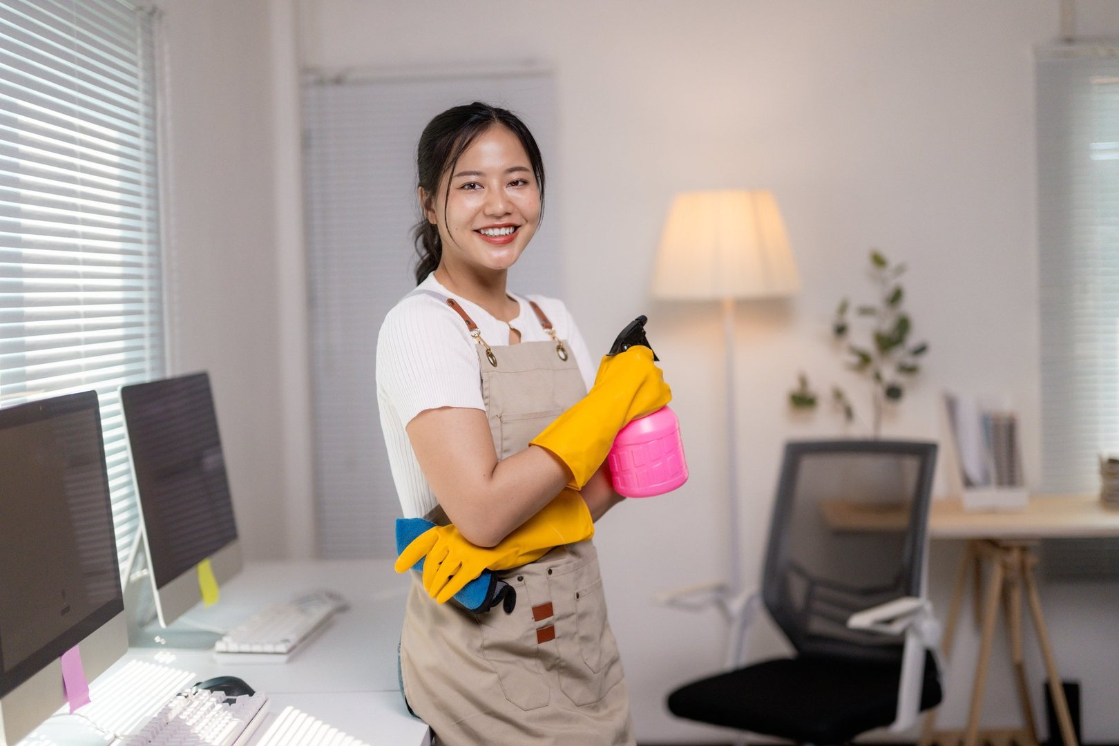 Smiling Asian female janitor holding cleaning supplies while working in a tidy office environment, showcasing professionalism and positivity