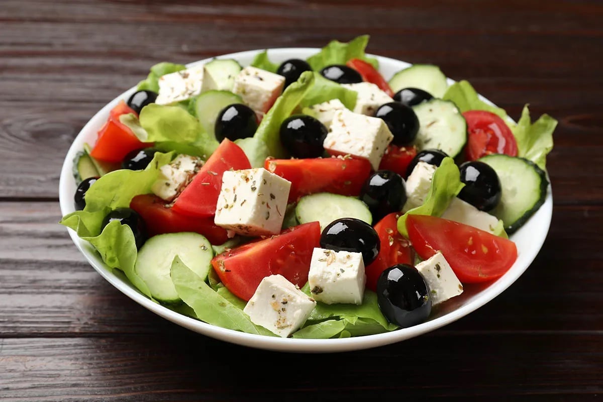 Bowl of Greek salad with lettuce, tomatoes, cucumbers, black olives, and feta cheese on a dark wooden table.