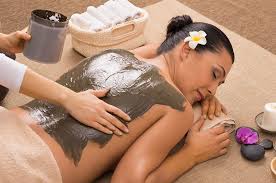 A woman receives a mud body treatment at a spa. She's lying on a massage table with a flower in her hair, next to a basket of towels.