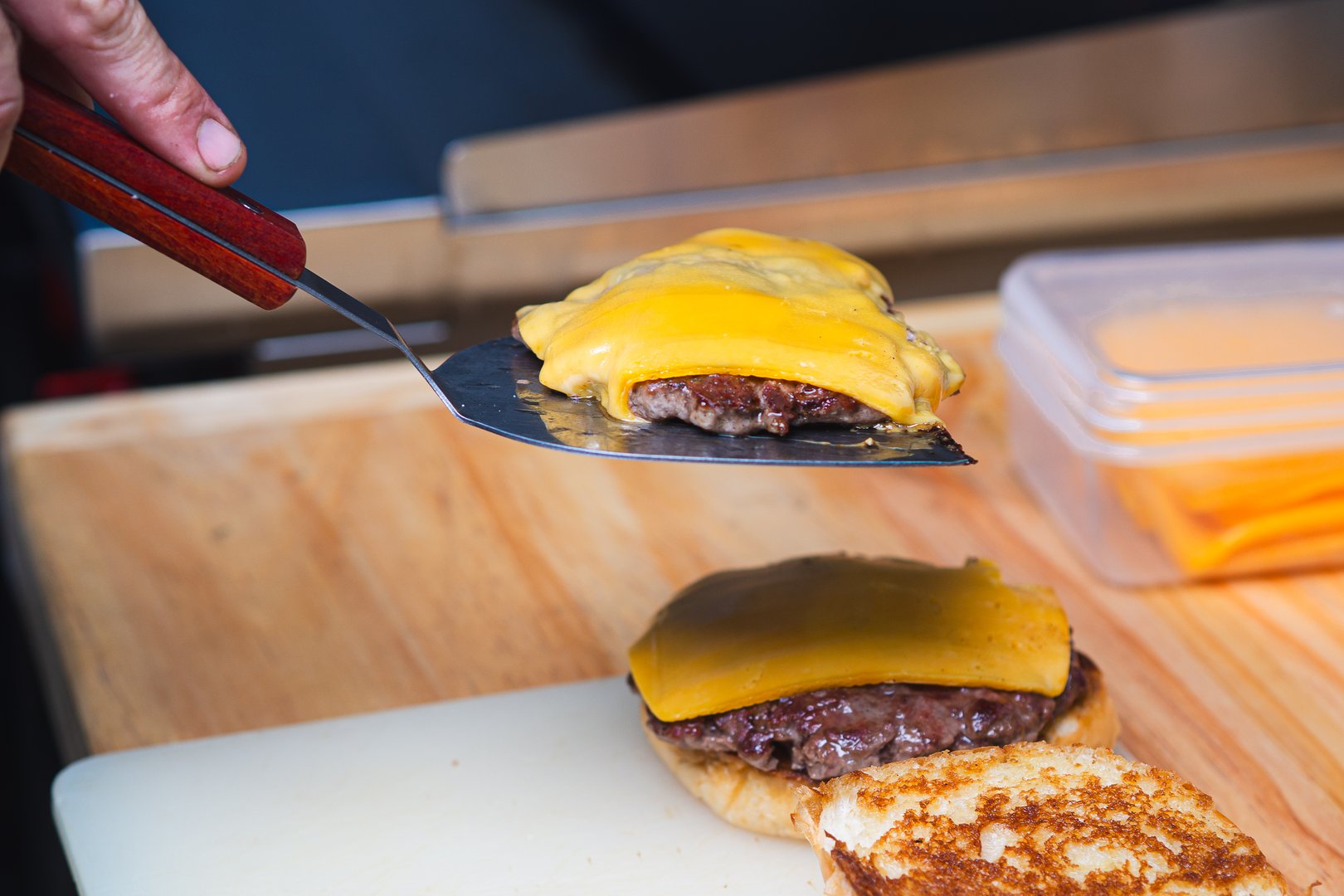 Grilled beef patty topped with melted cheddar cheese being lifted with spatula, ready to complete a delicious cheeseburger on toasted bun, perfect for fast food or barbecue themes.