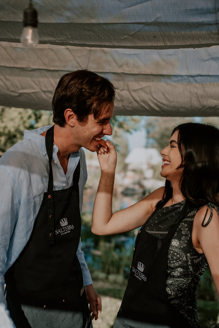 Two people wearing aprons smiling at each other, one feeding the other, outdoors under a canopy during daytime.