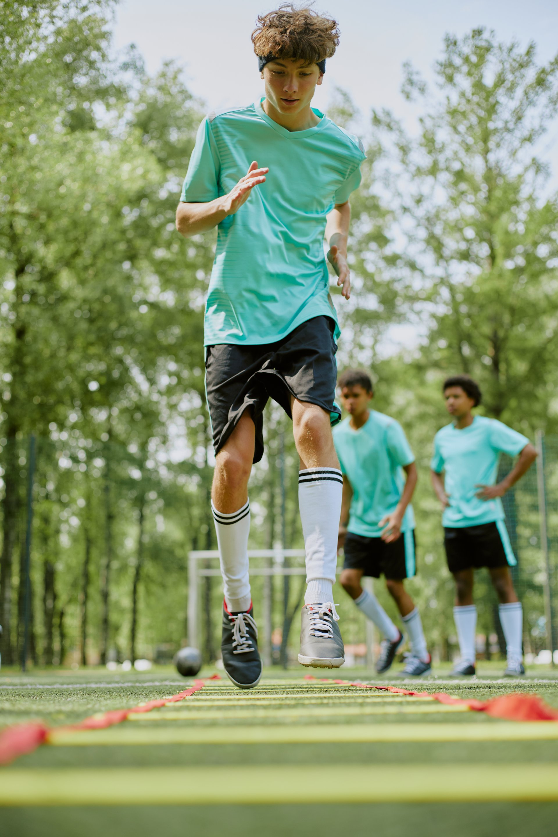 Teenage Caucasian boy training on soccer field performing agility ladder drill while two teenage boys standing in background watching practice session
